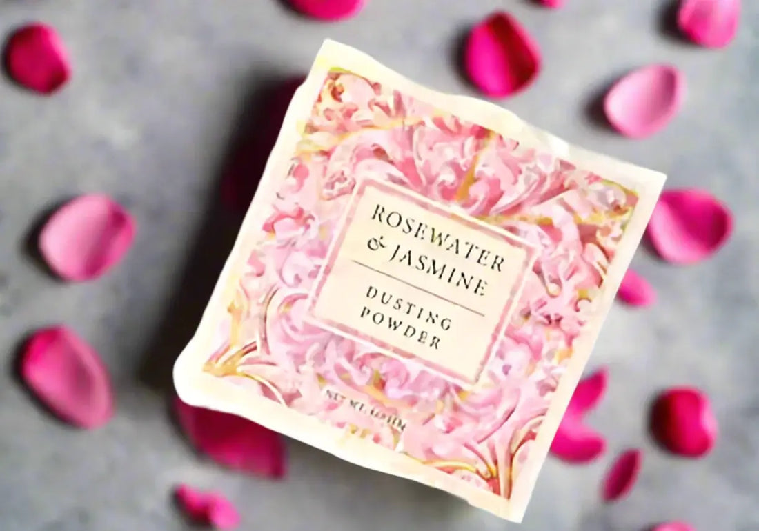 Botanical Rosewater and Jasmine Dusting Powder box surrounded by pink rose petals on a gray surface.